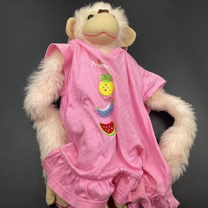 May include: A pink plush monkey wearing a pink dress with embroidered fruit designs. The dress has a ruffled hem and the text "mini me" is embroidered on the front.