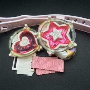 May include: A pink and white plastic toy set with a heart and star charm. The charms are attached to a pink strap and a white plastic base.