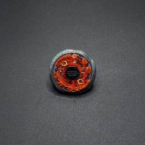 Może przedstawiać: Czarno-pomarańczowy Beyblade z przezroczystym, pomarańczowym plastikowym wierzchołkiem i czarną metalową podstawą. Wierzchołek ma wzór płomieni, a na podstawie widoczny jest napis "METAL FIGHT".