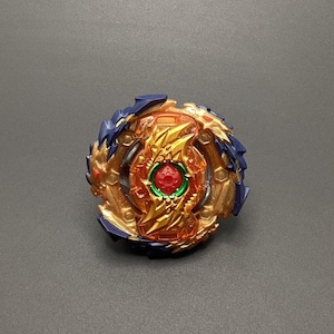 Puede incluir: Una peonza dorada y azul con un centro rojo y verde. La peonza tiene un diseño de dragón y está hecha de plástico.