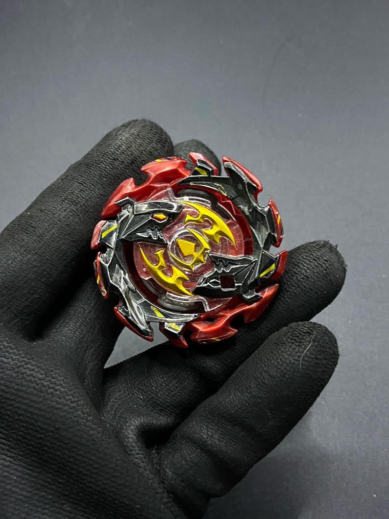 K&ouml;nnte beinhalten: Ein rotes und schwarzes Beyblade mit einem gelben und silbernen Design. Das Beyblade hat ein scharfes, kantiges Design mit einer durchsichtigen Plastikmitte.