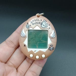 Puede incluir: Un dispositivo de mascota virtual Angel Gotch de color marrón con una pantalla que muestra un diseño de estrella. El dispositivo tiene botones blancos, detalles de alas de ángel y el texto "ANGEL GOTCH".