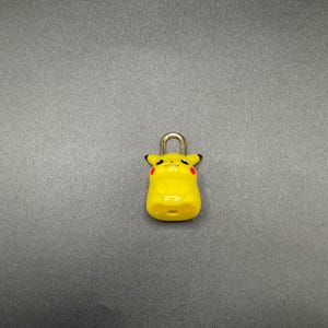 May include: A yellow Pikachu-shaped padlock with a silver-colored metal shackle. The padlock features black accents and red cheek markings, and is a collectible item.