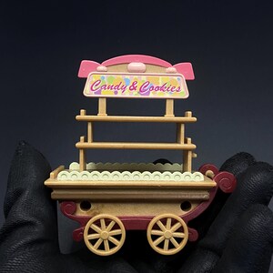 May include: A miniature candy and cookie cart with a pink and tan color scheme. The cart features a sign that reads "Candy & Cookies" and has two shelves. The cart has tan wheels and a red base.