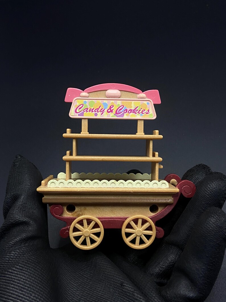 May include: A miniature candy and cookie cart with a pink and tan color scheme. The cart features a sign that reads "Candy & Cookies" and has two shelves. The cart has tan wheels and a red base.