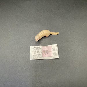Puede incluir: Una pequeña figura de hurón marrón claro con ojos rojos y una cola larga. La figura está sobre una superficie gris oscuro. Una pequeña etiqueta de papel con texto japonés y una ilustración de hurón está al lado.