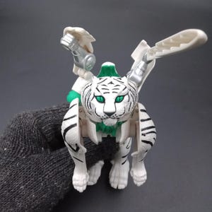 May include: A white and black tiger-like toy with green eyes and accents. The toy has articulated limbs and wings, with silver and white details. The toy is held by a person wearing a gray glove.