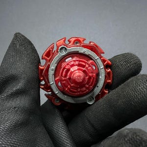K&ouml;nnte beinhalten: Rotes und silbernes Beyblade mit sternf&ouml;rmigem Design. Das Beyblade besteht aus Kunststoff und Metall.