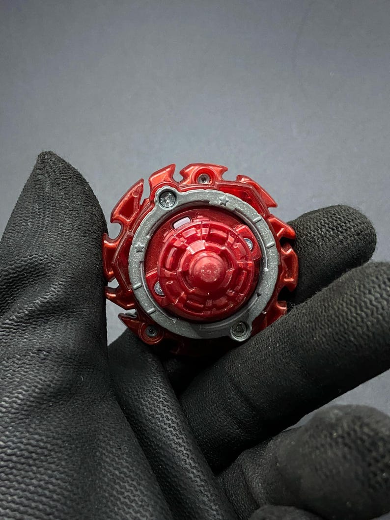 K&ouml;nnte beinhalten: Rotes und silbernes Beyblade mit sternf&ouml;rmigem Design. Das Beyblade besteht aus Kunststoff und Metall.