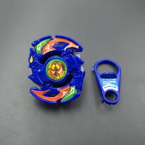 May include: A blue spinning top toy featuring orange and green accents on its circular frame, with a central circular emblem depicting a figure. Beside it lies a blue plastic tool with Japanese text and a white arrow printed on its surface.