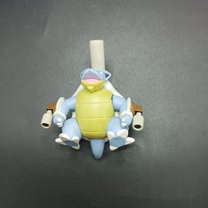 May include: A blue, yellow, and white Blastoise figurine with a light gray tube attached to its back. The figurine has a yellow shell, blue limbs, and white accents. The figure is set against a dark gray background.