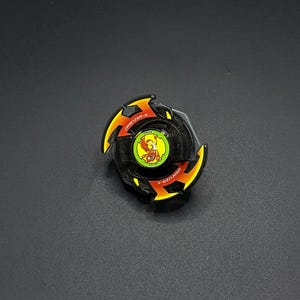 Pode incluir: Um brinquedo de pião preto e laranja com um emblema circular central num fundo amarelo com um gráfico de uma fénix vermelha. O anel exterior apresenta o texto "DRANZER-S" em letras brancas sobre detalhes laranja numa estrutura preta.