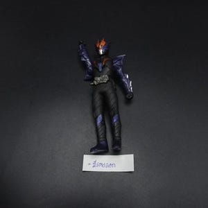 May include: A black and purple action figure with orange accents, standing on a white label with text. The figure is in a dynamic pose, with one arm raised. The figure has a detailed design, with armor-like elements.