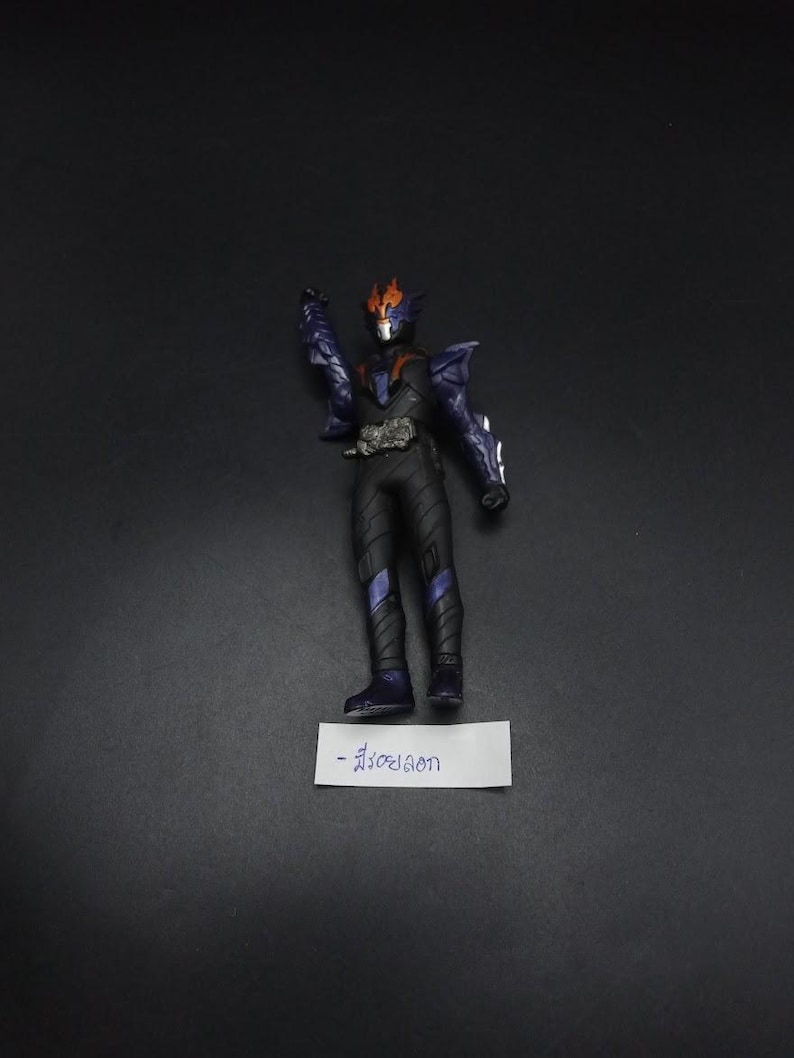 May include: A black and purple action figure with orange accents, standing on a white label with text. The figure is in a dynamic pose, with one arm raised. The figure has a detailed design, with armor-like elements.