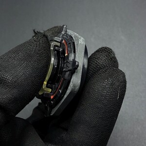 K&ouml;nnte beinhalten: Ein schwarzer und grauer Metall-Beyblade mit roten und goldenen Akzenten. Der Beyblade wird in einer behandschuhten Hand gehalten.