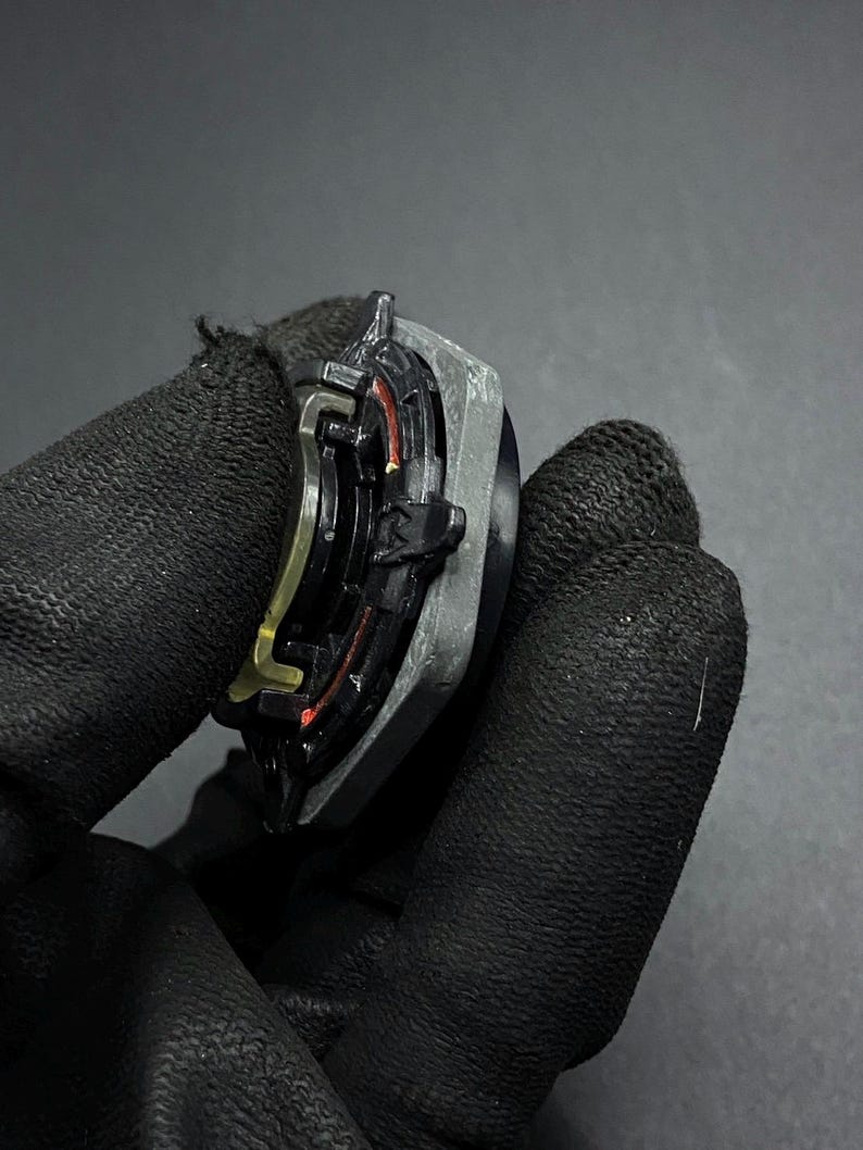 K&ouml;nnte beinhalten: Ein schwarzer und grauer Metall-Beyblade mit roten und goldenen Akzenten. Der Beyblade wird in einer behandschuhten Hand gehalten.