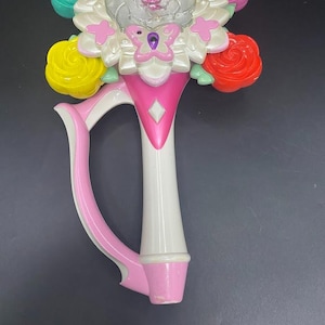 May include: A colorful toy wand with a flower-shaped top and a handle. The wand features a central flower with a pink center and clear petals, surrounded by smaller flowers in various colors, including pink, yellow, red, and teal. The handle is white and pink.