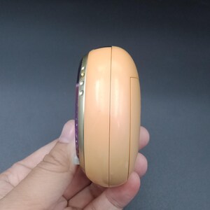 May include: A peach-colored, rounded electronic device with a gold-colored trim. The device has a small screen and buttons on the side. The device is held in a hand.