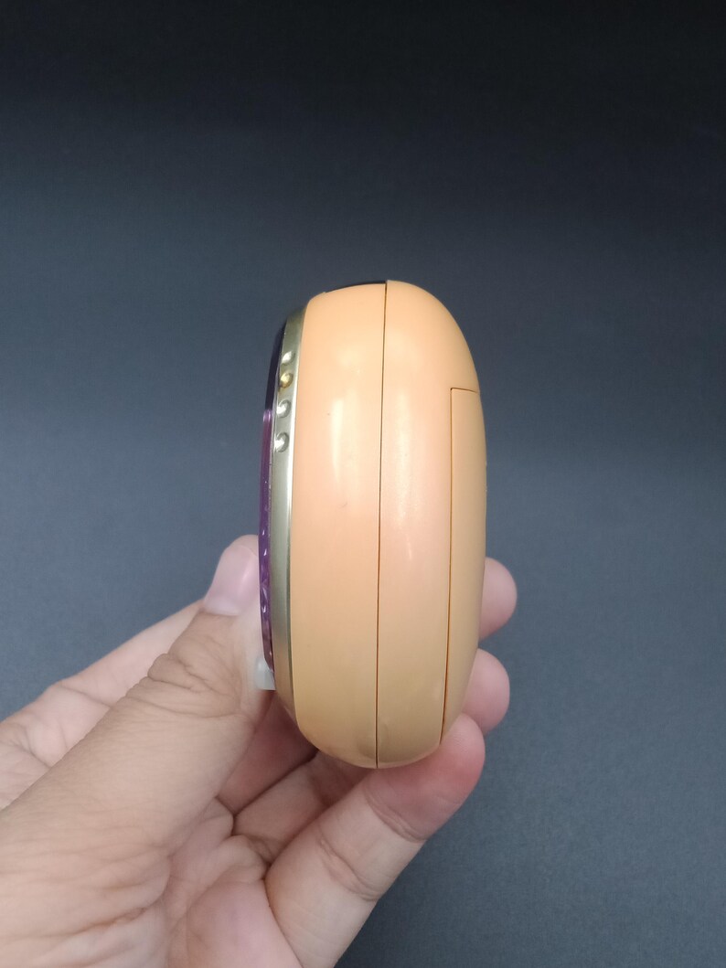 May include: A peach-colored, rounded electronic device with a gold-colored trim. The device has a small screen and buttons on the side. The device is held in a hand.