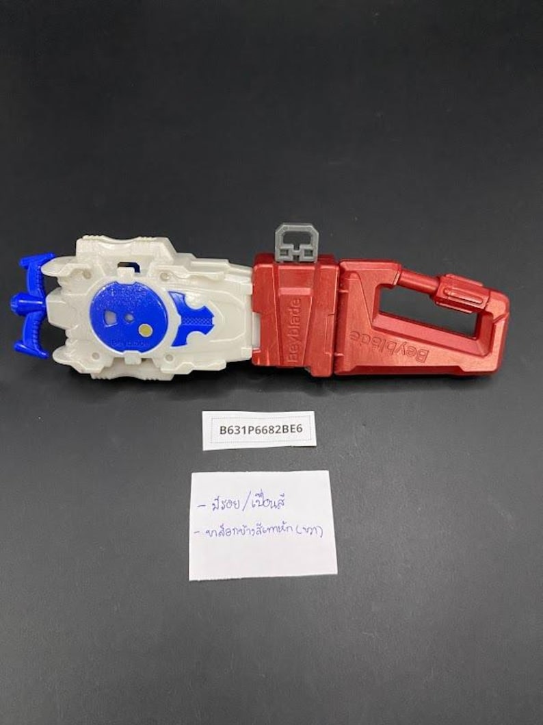 May include: A red and white Beyblade launcher toy with blue and white accents. The launcher has a handle and a trigger. The text "Beyblade" is visible on the side. The toy has the text "B631P6682BE6" on a sticker.