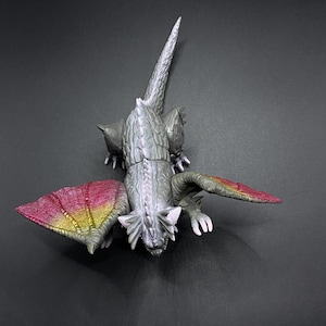 Peut inclure: Un jouet de dragon gris et blanc avec des ailes roses et jaunes. Le dragon est en position accroupie, les ailes déployées.