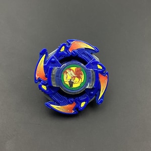 May include: Blue and red plastic Beyblade with a clear plastic center and a red and yellow phoenix design. The Beyblade has the words "Attack" and "Defense" printed on it.
