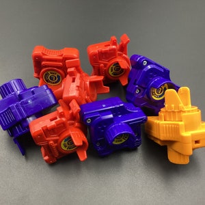 Può includere: Una collezione di sei trottole di plastica colorate. Le trottole sono in varie tonalità di blu, arancione e rosso. Ogni trottola ha un design circolare sulla parte superiore e un piccolo foro rotondo al centro.