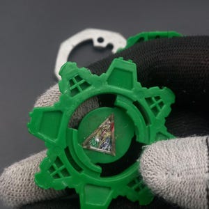 May include: A green plastic gear-shaped toy with a colorful triangular emblem in the center. The toy has a star-like design with multiple points. A white metal piece is visible in the background.