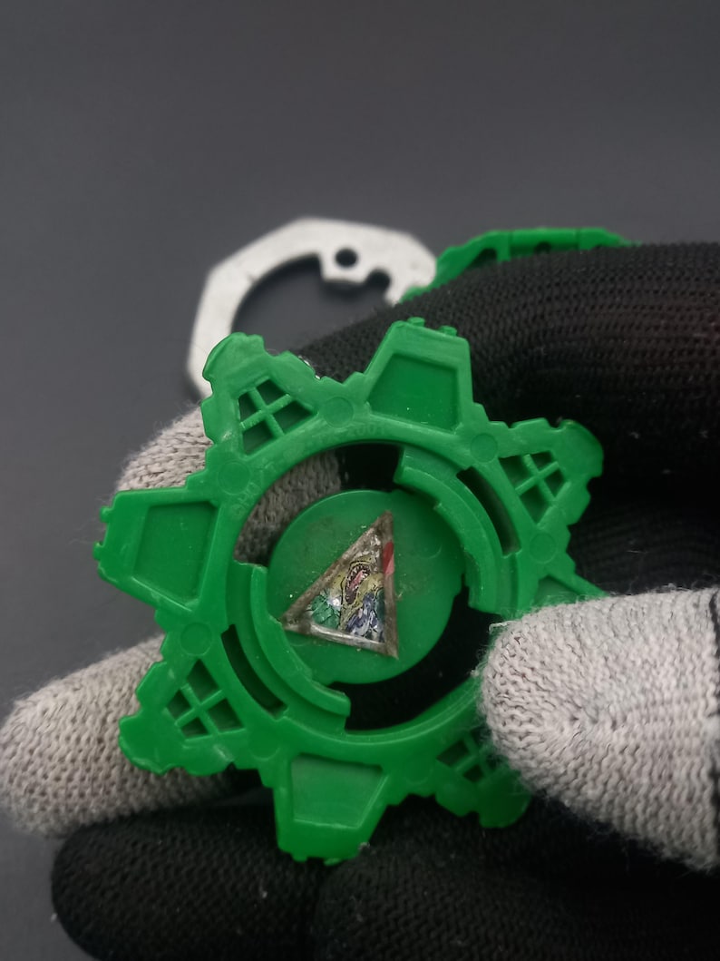 May include: A green plastic gear-shaped toy with a colorful triangular emblem in the center. The toy has a star-like design with multiple points. A white metal piece is visible in the background.