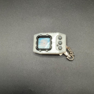 Puede incluir: Un juego digital portátil plateado con una pequeña pantalla que muestra una imagen pixelada. Tiene tres botones grises en el lado derecho y un llavero de metal. El dispositivo está sobre una superficie gris oscuro.