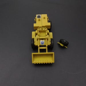 May include: A yellow toy construction vehicle with black wheels and a front scoop. The toy has a blue and white emblem on the front. A detached black tire is next to the vehicle. The toy is on a dark background.