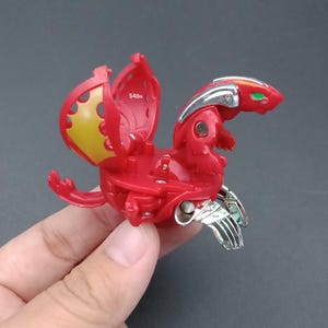 Può includere: Un giocattolo Bakugan rosso in stato parzialmente aperto, raffigurante una creatura simile a un drago con accenti metallici argentati sulla testa e sui piedi, dettagli gialli sulle ali interne e il testo 540G stampato sulla superficie rossa.