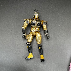 Könnte beinhalten: Gold-schwarze Roboter-Actionfigur mit Helm und Antenne. Die Figur hat bewegliche Gelenke und trägt einen gold-schwarzen Anzug mit gelben Akzenten an den Füßen.