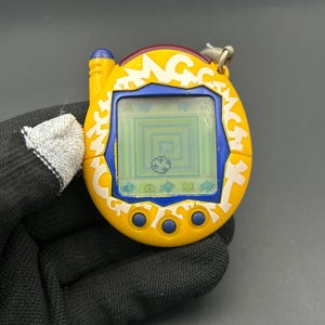 May include: A yellow and blue handheld digital pet game with a screen displaying a pixelated graphic. The device has a yellow casing with white text and blue buttons. A silver keychain is attached to the top.