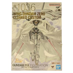 May include: A product box for the Wing Gundam Zero Cloaked Custom action figure, numbered 1036. The box features an illustration of a white robotic figure wearing a hooded cloak, surrounded by floating white feathers, with Bandai Namco branding.