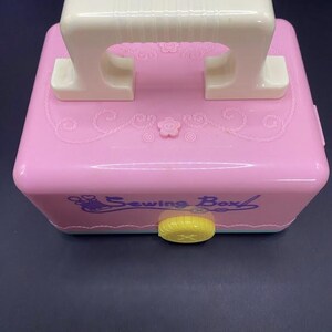 Puede incluir: Una caja de costura rosa con asa blanca y cierre amarillo. La caja tiene patrones florales decorativos y las palabras "Sewing Box" en cursiva. El asa mide aproximadamente 13 cm de largo.