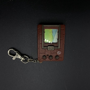 May include: A brown digital pet keychain with a screen displaying a green and blue landscape. The keychain has a silver metal clasp and four buttons.