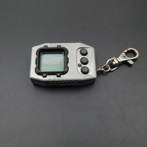 May include: A silver handheld digital pet game with a small screen displaying a pixelated landscape. It has three black buttons on the right and a keychain clip attached. The device is rectangular with a black border around the screen.