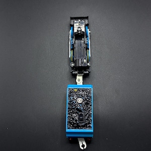 Könnte beinhalten: Eine blaue und schwarze Spielzeug-Lokomotive mit einer strukturierten schwarzen Front und einem blauen Sockel. Die Spielzeug-Lokomotive hat einen Haken an der Unterseite und ist vor einem dunklen Hintergrund platziert.