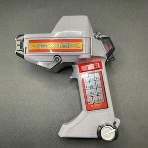 May include: A grey toy gun with red and yellow detailing. The toy gun features the text "GWGX" and a keypad with numbers and symbols. A white dial is located at the base of the handle.