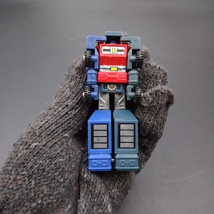 May include: A small, colorful toy robot with a red chest, blue legs, and gray arms. The toy has small wheels and rectangular details. The robot is held in a gray knitted glove.