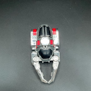 Könnte beinhalten: Ein silbernes und schwarzes Spielzeugfahrzeug mit roten Akzenten. Das Spielzeug hat einen zentralen schwarzen Cockpitbereich, ein großes zentrales Licht und zwei silberne Arme, die sich nach unten erstrecken. Das Fahrzeug ist futuristisch gestaltet.