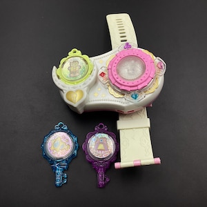 Peut inclure: Une montre-bracelet blanche et rose avec une bordure dorée et trois breloques clés colorées. Le cadran de la montre a une bordure rose et un centre transparent. Les breloques clés sont bleues, violettes et vertes avec des accents de paillettes.