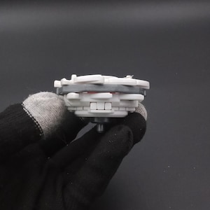 May include: A grey and white toy top with red detailing. The toy has a layered design and a central peg. The toy is held by a gloved hand against a dark background. This toy is designed for spinning.