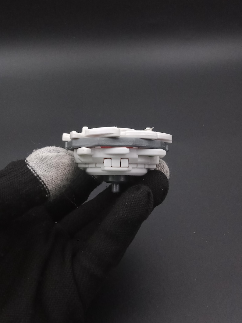 May include: A grey and white toy top with red detailing. The toy has a layered design and a central peg. The toy is held by a gloved hand against a dark background. This toy is designed for spinning.