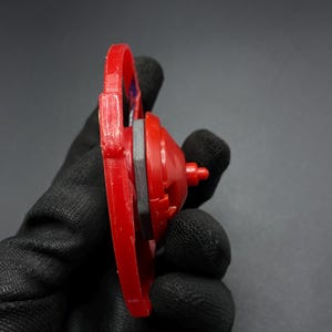 May include: Red and black plastic toy top with a silver metal piece in the center. The top is held in a gloved hand.