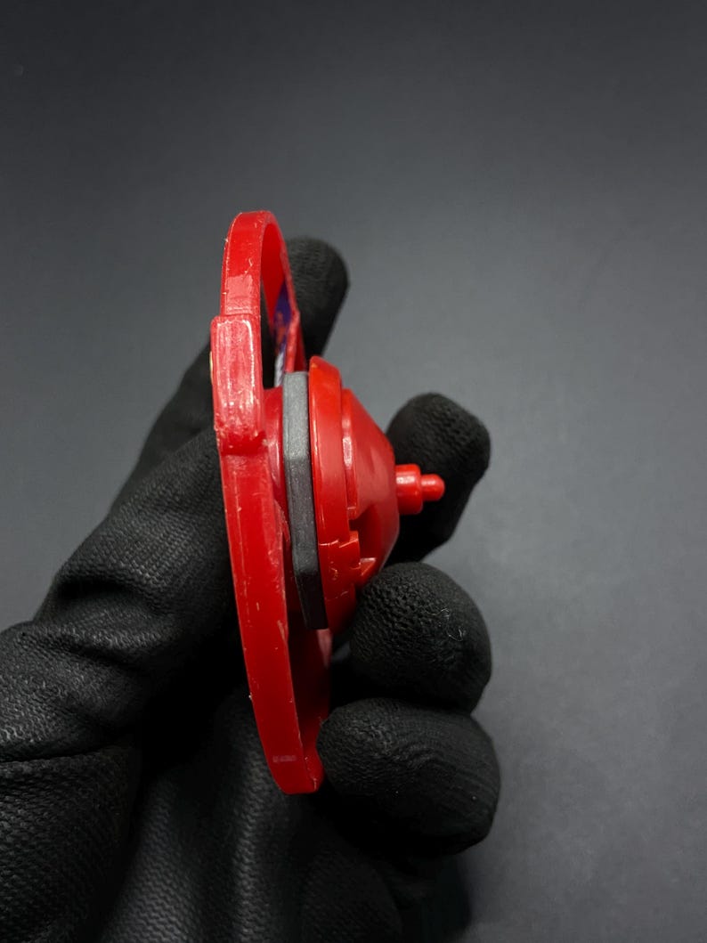 May include: Red and black plastic toy top with a silver metal piece in the center. The top is held in a gloved hand.