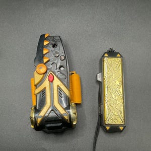 Puede incluir: Dos accesorios de juguete: un dispositivo negro y dorado con detalles en naranja y un colgante rectangular dorado con un cordón negro. El dispositivo tiene un diseño complejo con una gema roja y una serie de botones. El colgante presenta un patrón ornamentado.