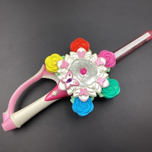 以下が含まれることがあります： ピンクと白の玩具の剣で、銀色のセンターピースと蝶のデザインが施されています。剣は、ピンク、赤、黄、青、緑のカラフルな花で飾られています。