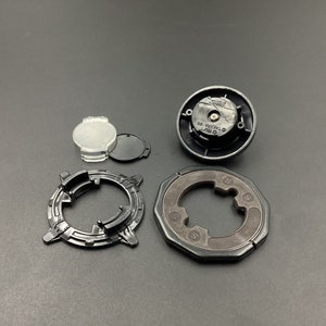 May include: A set of black plastic Beyblade parts, including a clear plastic piece, a black plastic piece with a screw, a black plastic piece with a circular design, and a black plastic piece with a hexagonal design.